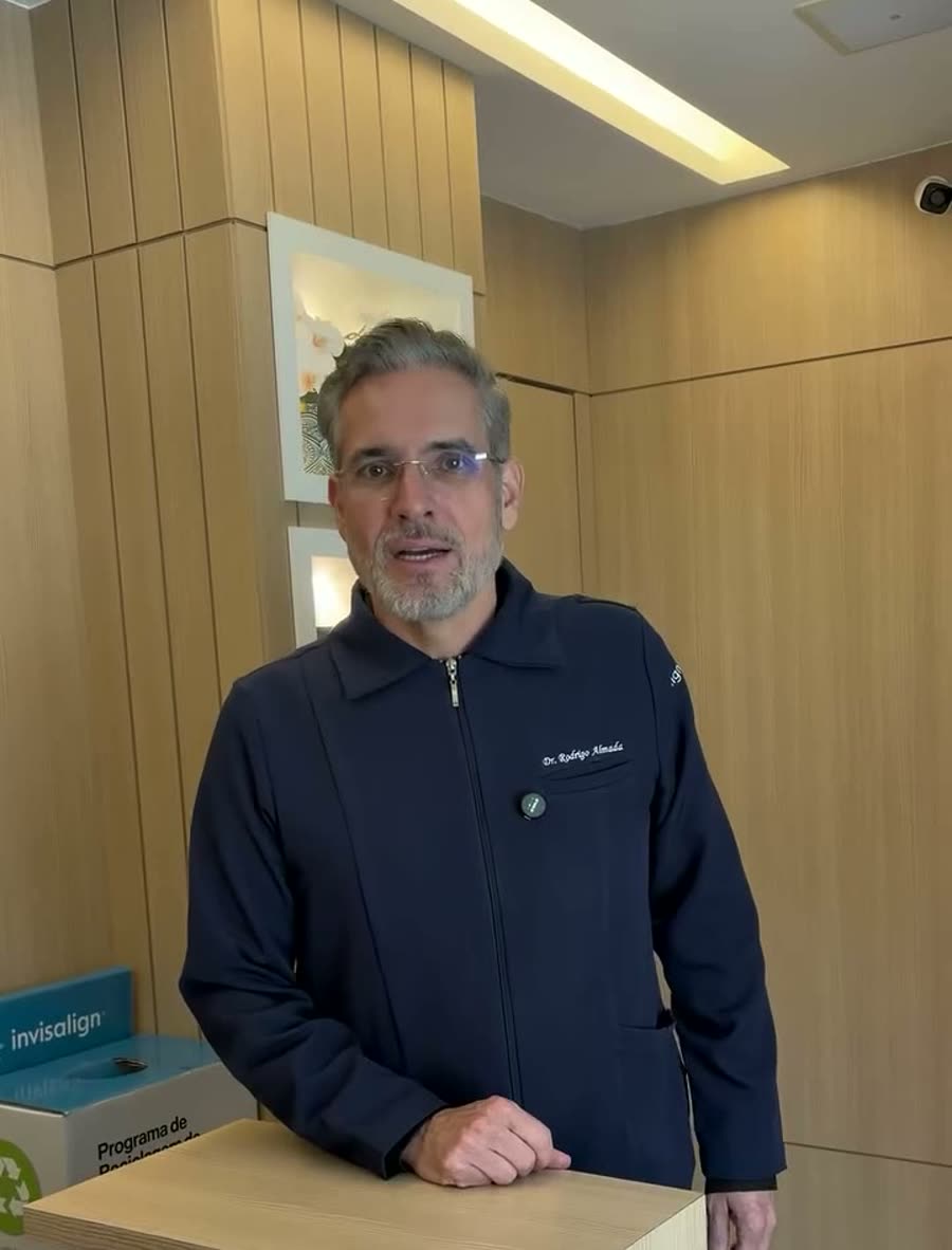 Dr. Rodrigo Almada em pé na clínica, diante do painel de madeira da recepção